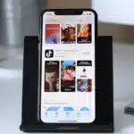 Un smartphone sur un support noir affiche la page de l'application TikTok dans l'App Store, avec des images, le logo TikTok et le bouton Ouvrir. Un clavier et une partie d'un écran sont visibles en arrière-plan.