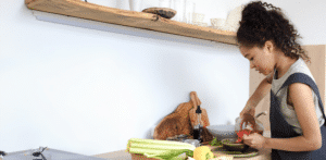 Une femme prépare des aliments dans une cuisine, versant de la sauce dans un bol. Des légumes frais, dont du céleri et du citron, sont posés sur le comptoir. Des étagères contenant de la vaisselle sont visibles au-dessus d'elle.