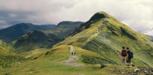 Des randonneurs marchent le long d'un sentier de montagne sinueux entouré de collines et de sommets verdoyants sous un ciel nuageux, d'autres randonneurs étant visibles plus loin sur le chemin.