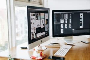 Deux écrans d'ordinateur posés sur un bureau en bois affichent des travaux de conception graphique. À proximité, un clavier, une souris, un smartphone, un carnet de notes, un stylo et une figurine de chat ondulant en céramique complètent la scène, offrant ainsi un aperçu de notre monde créatif : Qui sommes-nous ?.