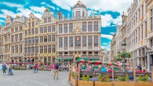 Des cafés en plein air sous des parasols colorés remplissent une place animée de Paris, entourée de bâtiments historiques ornés ; des professionnels des agences de presse se mêlent aux gens qui dînent et se promènent sous un ciel partiellement nuageux.
