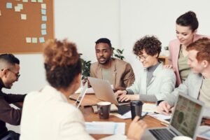 Un groupe de cinq personnes est assis et discute autour d'une table avec des ordinateurs portables, des documents et des tasses de café, collaborant dans un bureau lumineux de Bruxelles. Une personne semble parler tandis que les autres écoutent et sourient à leurs missions d'agence d'influence.