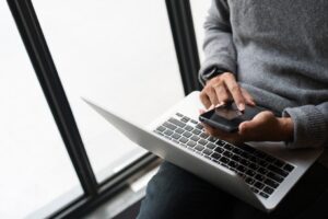 Personne vêtue d'un pull gris, assise près d'une fenêtre à Lille, utilisant un smartphone d'une main et posant un ordinateur portable - peut-être en train de travailler à distance pour des agences de communication. Seuls les mains et le torse sont visibles.