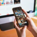 Une personne tenant un smartphone et naviguant dans une application de galerie de photos, avec un écran d'ordinateur affichant des images en arrière-plan sur un bureau en bois - des outils parfaits pour animer une communauté autour d'un contenu visuel.