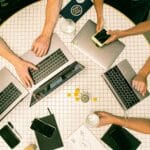 Vue de haut en bas de quatre personnes travaillant autour d'une table ronde - idéale pour les agences de marketing B2B - avec des ordinateurs portables, des smartphones, des carnets de notes, des boissons et des pièces de monnaie éparpillés sur une surface carrelée blanche.