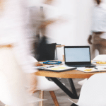 Une table ronde avec un ordinateur portable, des documents et des collations dans un bureau moderne d'une agence de communication B2B. Plusieurs personnes passent, créant un effet de flou de mouvement, tandis que des tableaux et des graphiques sont visibles sur un tableau à l'arrière-plan.