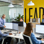 Dans un bureau moderne dont le mur jaune vif affiche le mot LEADS, un groupe de cinq personnes travaille sur des ordinateurs et collabore sur des générations de leads. Une personne se tient debout, pointant un écran, tandis que les autres sont assis et discutent.