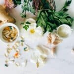 Sur une surface en marbre blanc, on trouve du gingembre frais, des herbes, de l'ail, une fleur blanche et des bols contenant des gélules et des comprimés, ce qui est idéal pour commercialiser des concepts nutritionnels. Quelques pilules et gousses d'ail sont éparpillées.