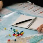 Une personne tient une loupe au-dessus d'une carte du monde parsemée d'épingles à nourrice colorées. Un carnet à spirales ouvert et un stylo reposent sur la carte, évoquant la préparation d'un voyage ou une recherche, peut-être par une agence de communication touristique.