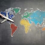 Une main tient une maquette d'avion au-dessus d'une carte du monde dessinée à la craie - les continents sont délimités en blanc et colorés en bleu, jaune, vert et rouge - symbolisant la façon dont la communication numérique révolutionne le tourisme dans le monde entier.