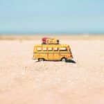 Une petite camionnette jaune avec des bagages sur le toit est posée sur un sol sablonneux, évoquant l'esprit d'aventure touristique sous un ciel bleu clair et un horizon lointain.