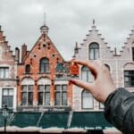 Une main tient une petite maquette souvenir de bâtiments colorés à pignons en escalier, correspondant aux véritables bâtiments historiques de Bruges, en Belgique, sous un ciel nuageux - une inspiration parfaite pour une campagne de l'agence de communication tourisme Belgique.