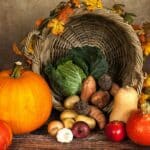 Une corne d'abondance en osier, parfaite pour une campagne de marketing nutritionnel, déborde de légumes verts, de pommes de terre, d'ignames, et est entourée de citrouilles, de courges, d'ail, de pommes et de feuilles d'automne sur une surface en bois rustique.