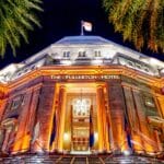 Le bâtiment de l'hôtel Fullerton de nuit, illuminé de lumières orange et blanches, vu en contre-plongée, avec des feuilles de palmier encadrant la scène - une vue qui inspire toute agence de communication spécialiste hôtellerie en Belgique. Les drapeaux sont affichés au-dessus de l'entrée.