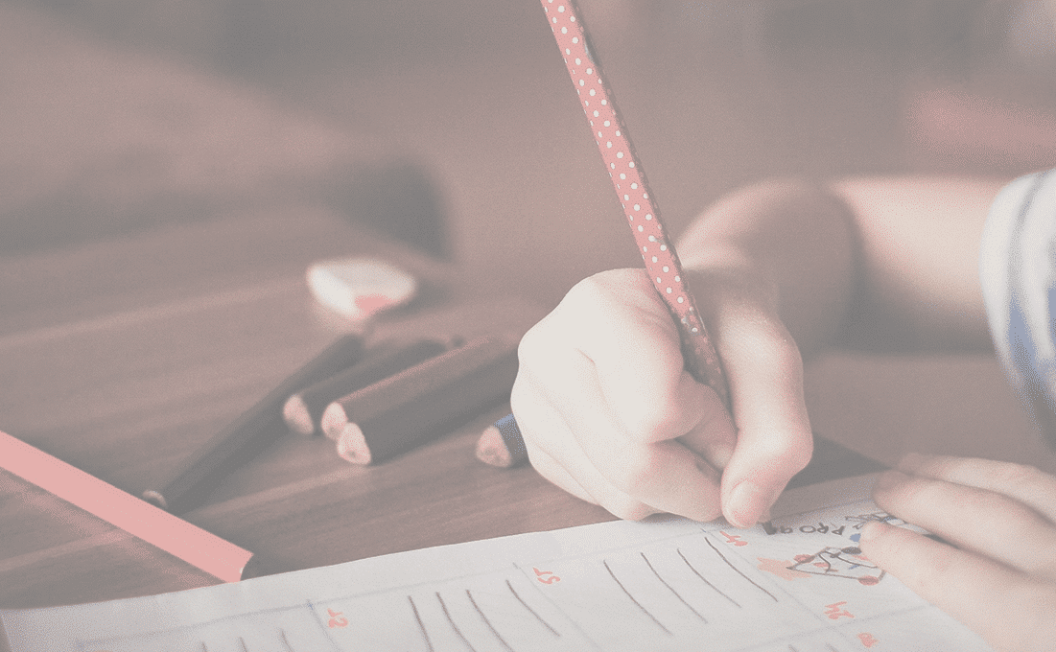 Un enfant s'adonne à l'éducation en écrivant sur une feuille de travail avec un crayon rouge, sur une table en bois, entouré de crayons de couleur et d'une gomme. L'accent est mis sur la main de l'enfant et sur la feuille.