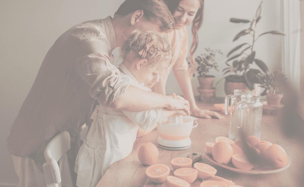 Une famille se tient autour d'une table de cuisine, un enfant pressant des oranges dans un bol tandis que deux adultes l'aident. La table est recouverte d'oranges coupées en deux et la lumière du soleil pénètre par une fenêtre voisine.