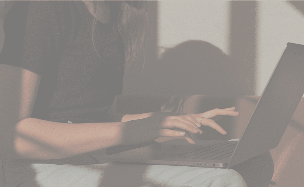 Une personne assise sur un canapé, tapant sur un ordinateur portable. La lumière du soleil projette des ombres sur ses bras et sur le clavier alors qu'elle travaille sur son projet de média. Seuls le haut du corps, les mains et l'ordinateur portable sont visibles.